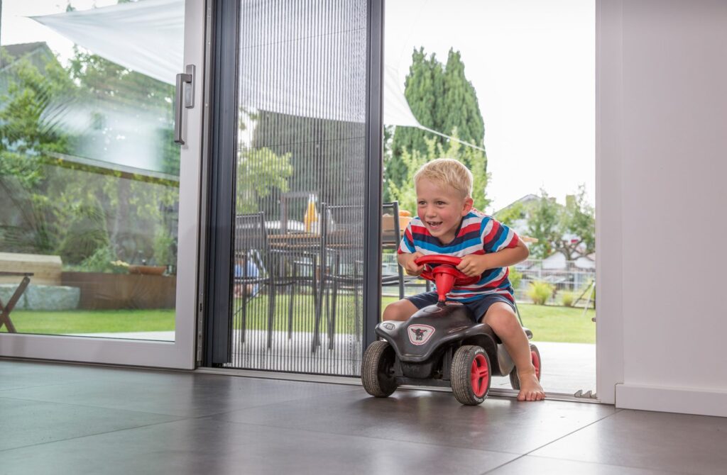 Schwellenfreie Insektenschutz-Plisseetür – Kind fährt mit Bobbycar hindurch